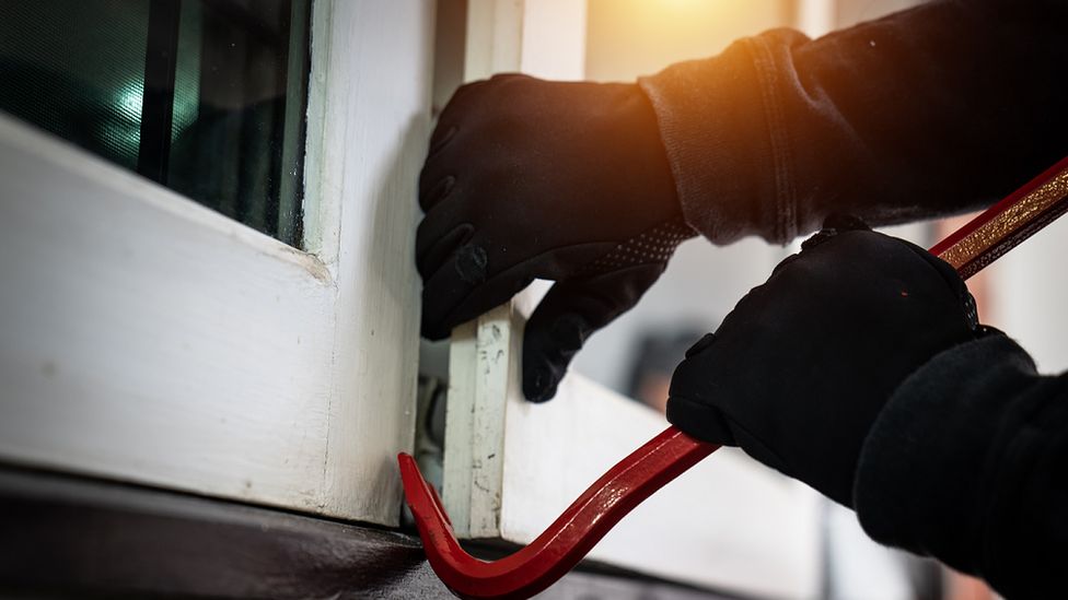 A stock image showing a person in gloves using a crowbar to open a window to gain access to a home