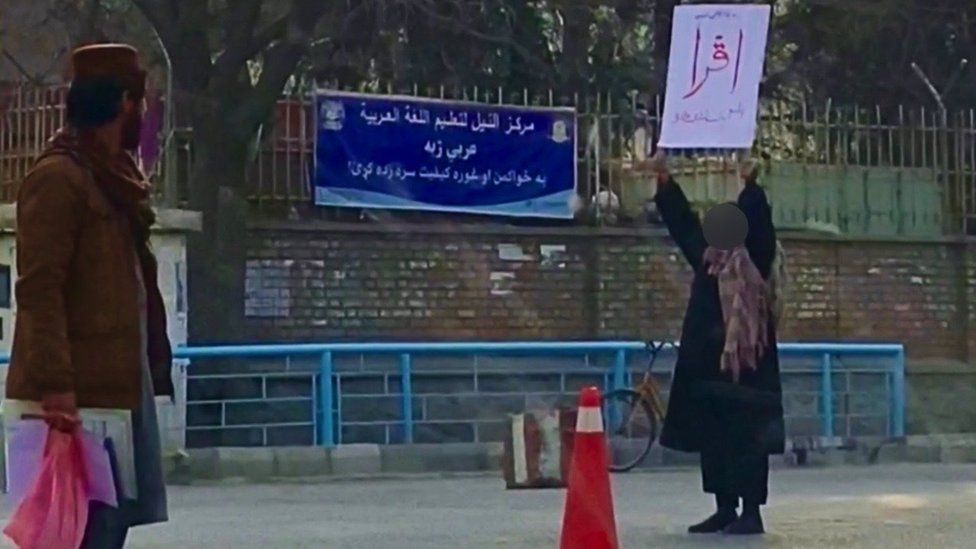 An onlooker watches Adela holding a placard