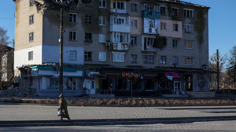 A woman walks across a road towards a housing block damaged by the war in Ukraine. Windows are smashed and masonry is falling away.