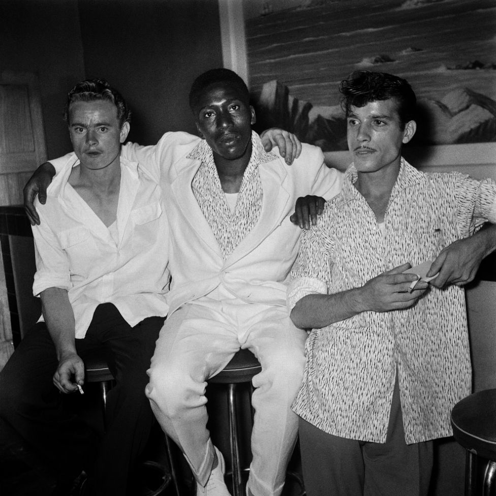 Three men dressed in loose shirts and holding cigarettes stand by the bar.