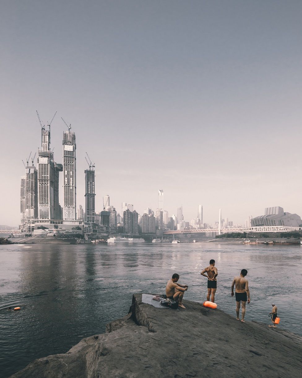 Buildings being constructed in the distance with swimmers next to a river in the foreground