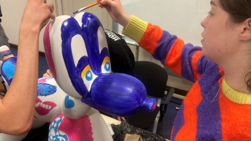 Balloon dog sculpture with a blue face and different colours across it's body being painted by two people