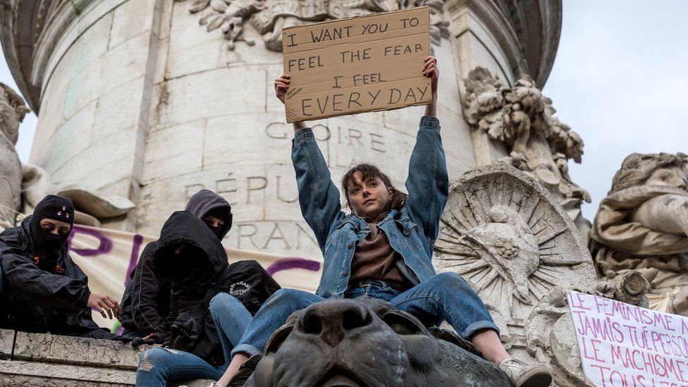 A young woman who has climbed one of of Paris's monuments holds aloft a sign in English, reading: "I want you to feel the fear I feel every day", as masked demonstrators sit nearby