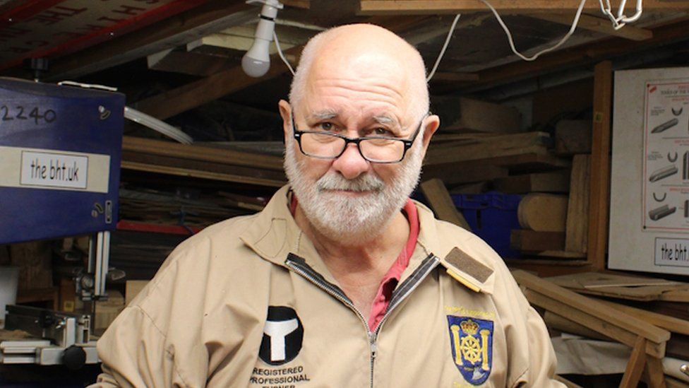 Wood turner John Boyne-Aitken with a short white beard and black-rimmed glasses, standing in a room full of wood.