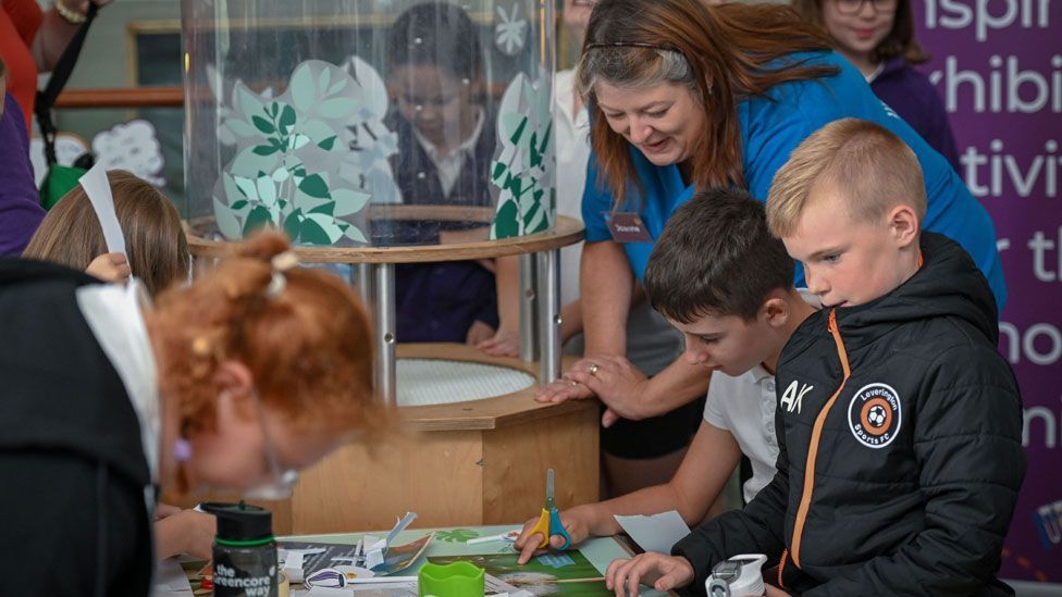 A woman in a blue polo shirt showing two boys and a girl science material 