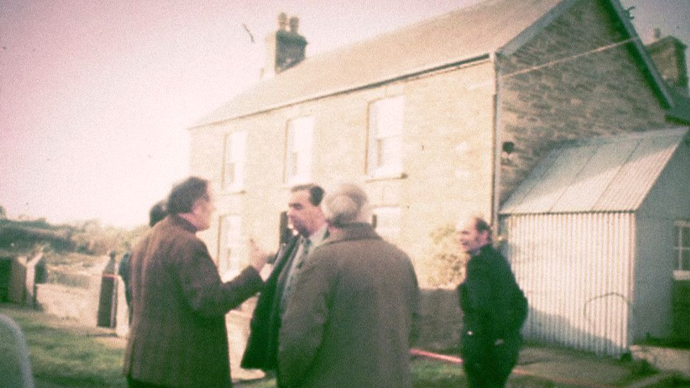 A still from archive footage of police officers outside Ffynon Samson farmhouse in December 1976. DCS Pat Malloy is stood in the centre of the group, wearing a suit and talking to his colleagues, as a uniformed officer looks on.