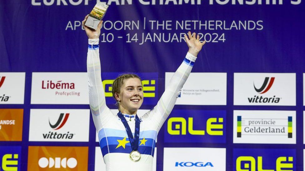 Winner Emma Finucane (GBR) with the gold medal during the ceremony of the final sprint on the third day of the European Track Cycling Championships in the Apeldoorn Omnisportcentrum. Emma is a 21-year-old white woman with blonde hair which she wears braided. She is dressed in a white cycling jersey with blue stripes across her chest and wrists. She holds a trophy in her right hand, both arms aloft, and smiles. She has a medal around her neck and is pictured in front of event hoarding for the cycling championships.