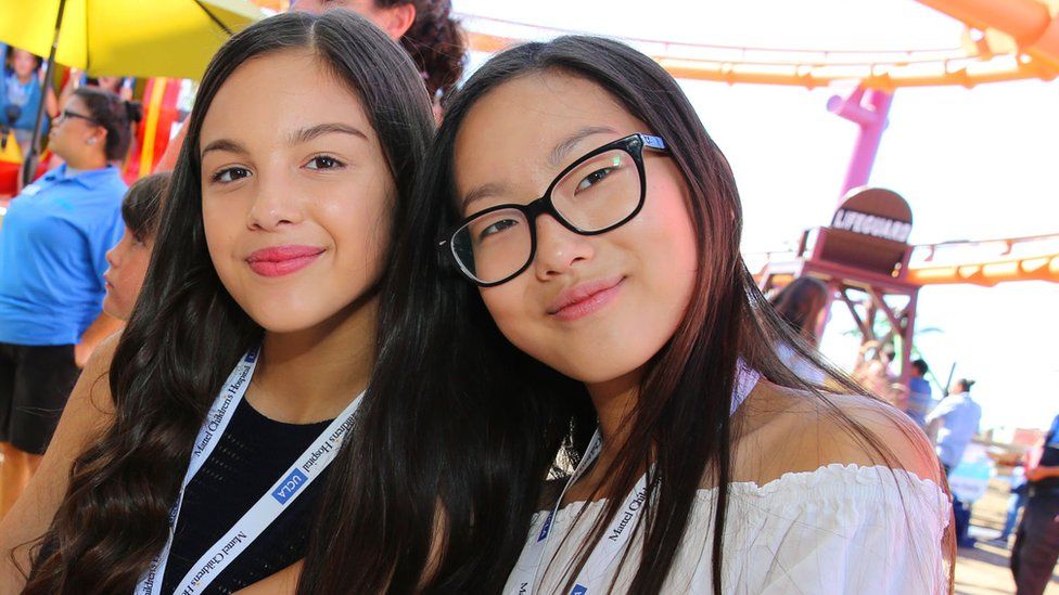 Olivia Rodrigo and Madison Hu in Santa Monica in 2016. The girls are in their early teens - Olivia, on the left, has long dark hair worn loose and wears a sleeveless black top. Monica also has long dark hair, she wears black square-rimmed glasses and a white off-the-shoulder blouse and leans into her friend. They both wear lanyards and are pictured outside at an amusement park,