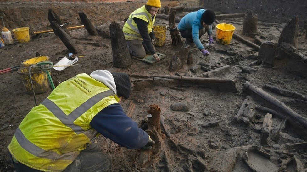 Three archaeologists in high visibility jackets excavating among mud and burnt timber remains