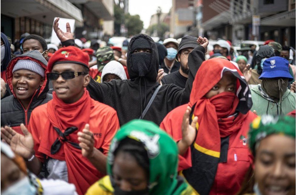 Students wearing red and black protest. One person in the middle is wearing a black hoodie and face mask.