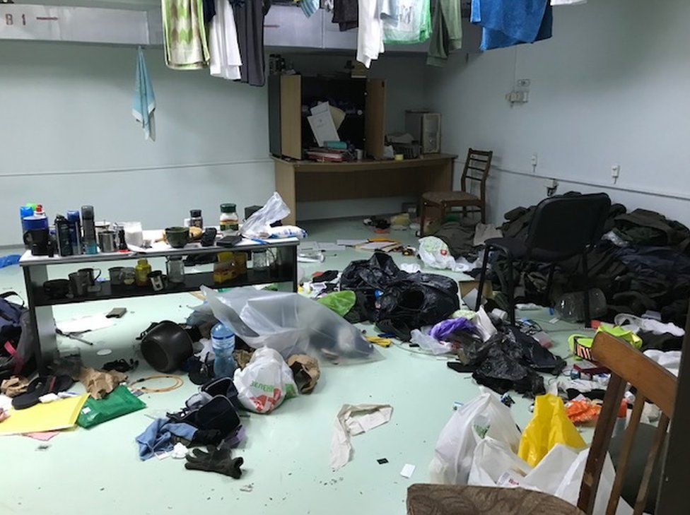 A chaotic room with a pale green floor covered with items like clothes, mugs, chairs, plastic bags