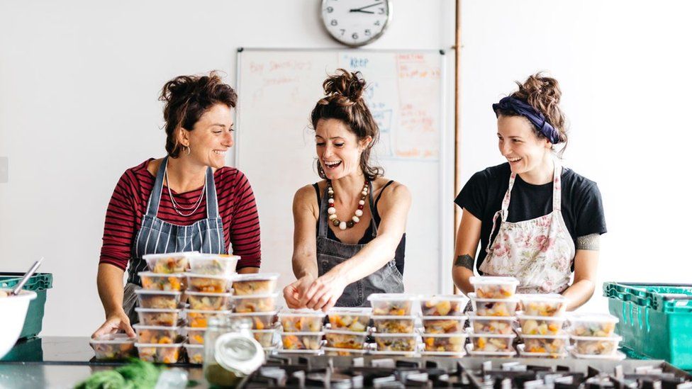 Three women stood making meals and packing them into containers