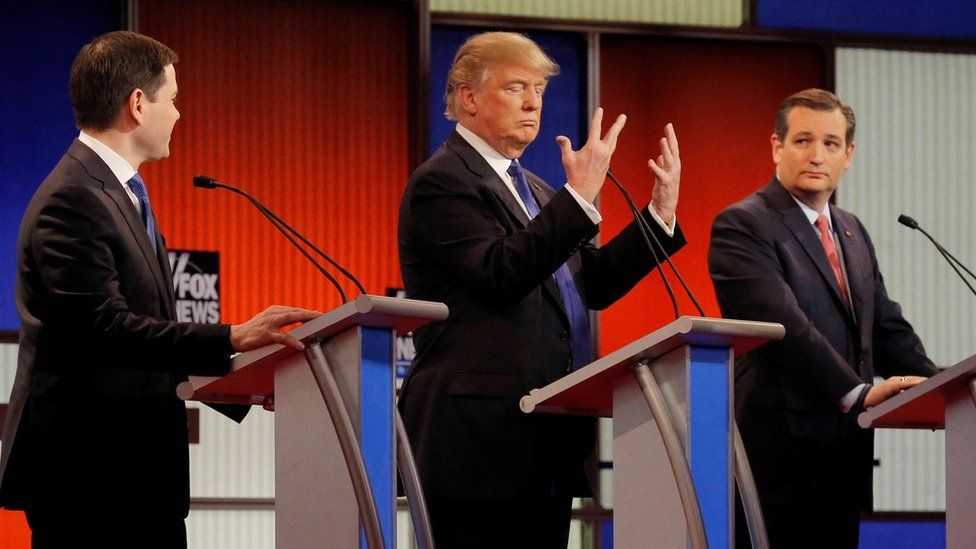 Trump looks at his hands and stands behind a podium. Ted Cruz stands to his right and Marco Rubio to his left.
