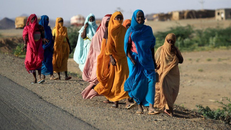 A group of women in colourful clothing walking in a line on the side of a road. They are on a dirt pavement. They are wearing traditional long dresses with hijabs. Their surroundings look rural.