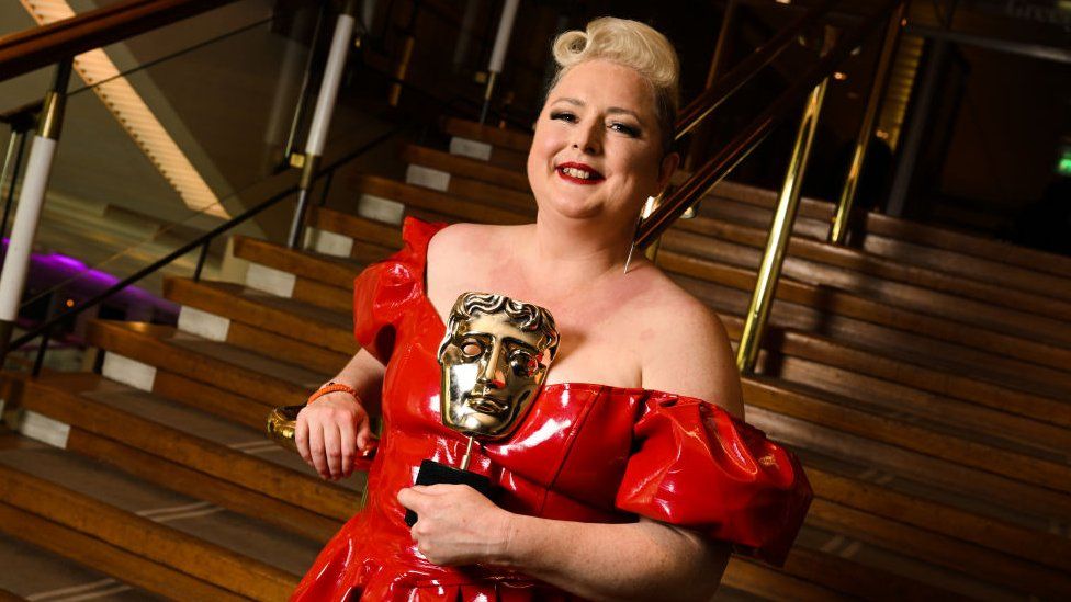 Siobhán McSweeney poses backstage with the Female Performance in a Comedy Programme Award for 'Derry Girls' at the 2023 BAFTA Television Awards. Siobhán is a white woman in her 40s, she has short blonde hair curled on top of her head and wears a bright red latex dress which she has matched with red lipstick. She holds a bronze BAFTA trophy and smiles at the camera. She is pictured in front of some stairs leaning on a gold hand rail