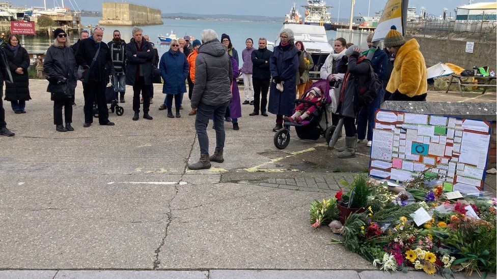 People standing at Portland Port next to a pile of flowers