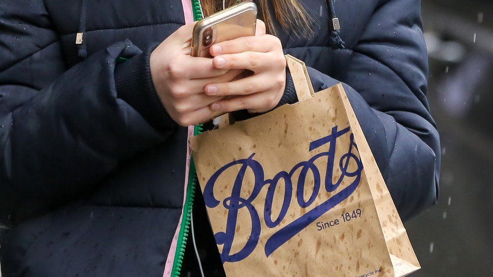 A woman's hands are seen holding a Boots paper bag and a mobile phone