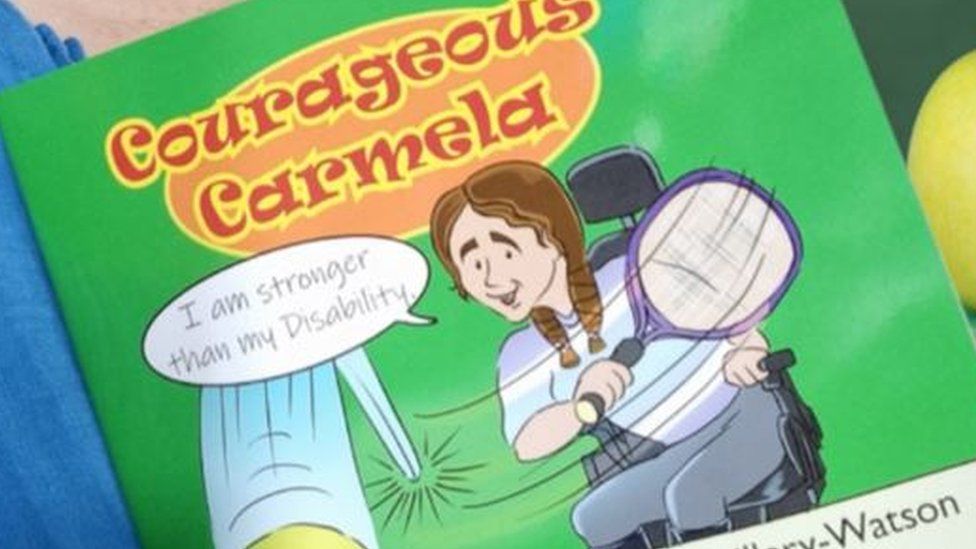 Lucy holds the book, which is green with a cartoon of Carmela in a wheelchair playing tennis saying 'I'm stronger than my disability'