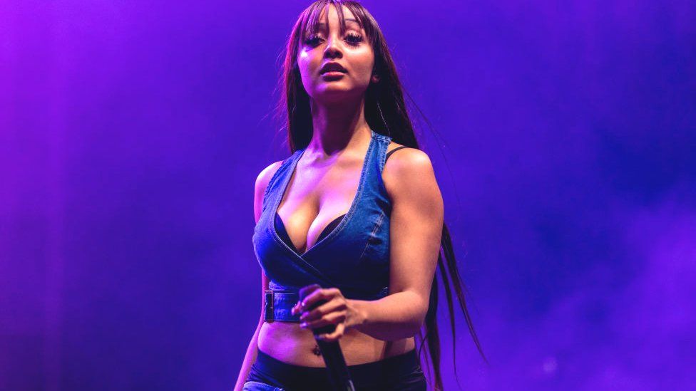 PinkPantheress performs at Camp Flog Gnaw Carnival in LA in November 2023. She has long brown hair down her back and a fringe just past her eyebrows. She wears a cropped denim waist coat and holds a microphone in her left hand. She is lit by purple stage lighting