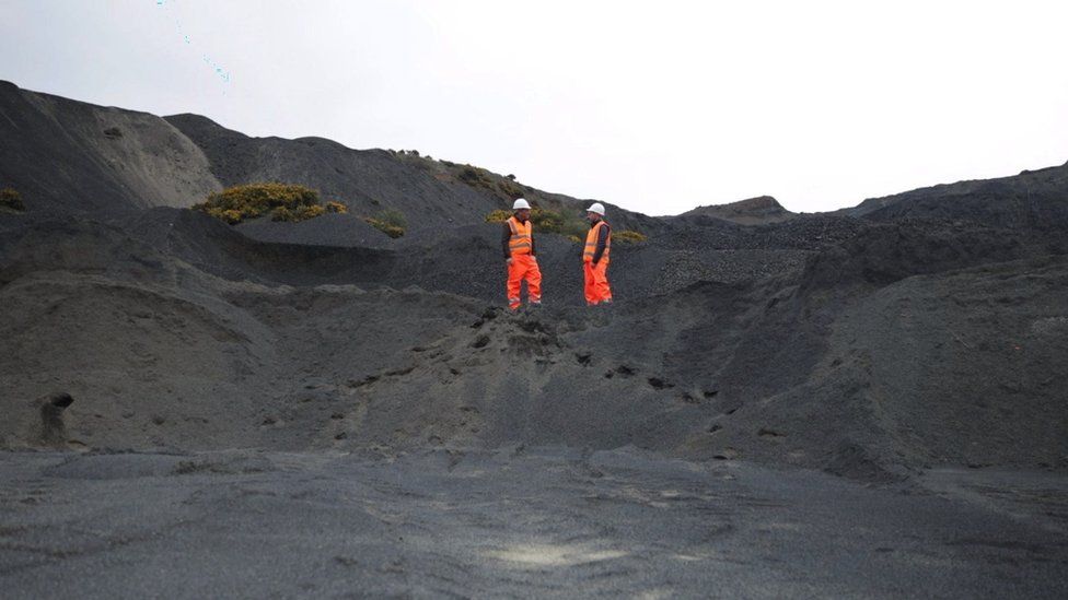 two men in orange hi-vis talk amongst hills of black rock
