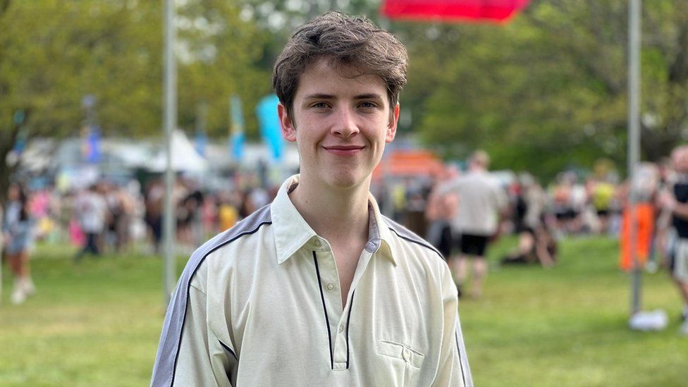 Luke Smith smiling for the camera at Radio 1's Big Weekend festival with people in the background. He is wearing a light coloured polo shirt