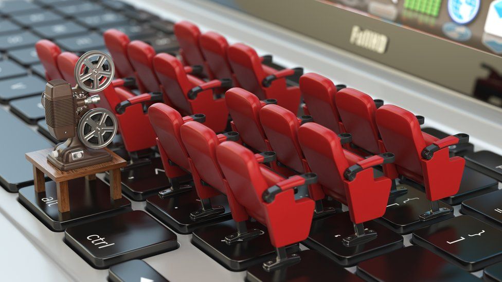 Model cinema seats are arranged in neat little rows on a laptop keyboard in this staged photo, while behind them, a model old-style film projector is perched, to illustrate the laptop screen being like a cinema
