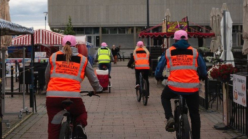 Four people in high-vis vests cycle in middle of market place and cafe area in Oulu