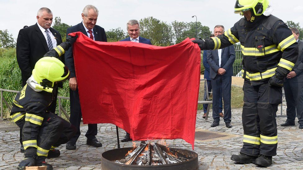 Czech politicians, including President Zeman, hold the giant boxer shorts aloft as two firefighters in full protective gear prepare to burn it in a small fire pit in front of him