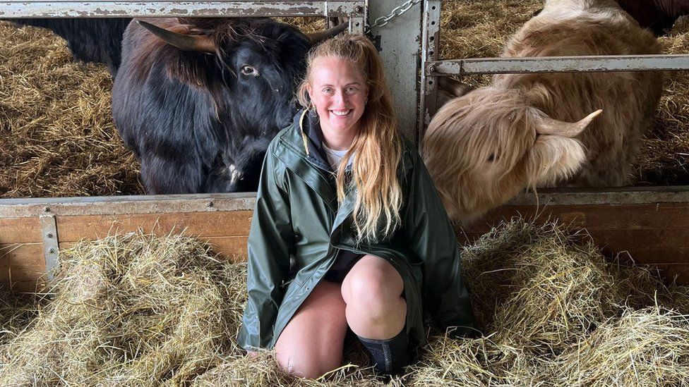 Izzi Rainey - a blonde woman in her early 30s kneels in a barn in front of two of her cattle, one black and one blonde. Izzi s a while woman and her hair, tied in a pony tail, is over her shoulder reaching down to her middle. She wears a dark green rain mac and knee high boots and kneels amongst some hay - both cows are taking a keen interest in Izzi.