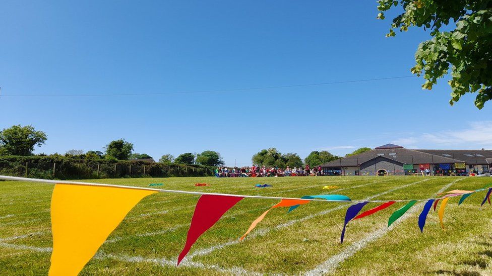 A string of coloured bunting runs along the top of a field marked out for a sporting event. In the background a large number of people have gathered around benches with bright blue sky above