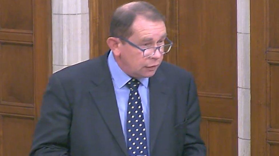 Man with short brown hair and glasses standing in front of wood panelling in Westminster Hall