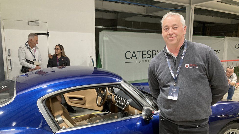 Man with short white hair wearing lanyard standing next to low blue car