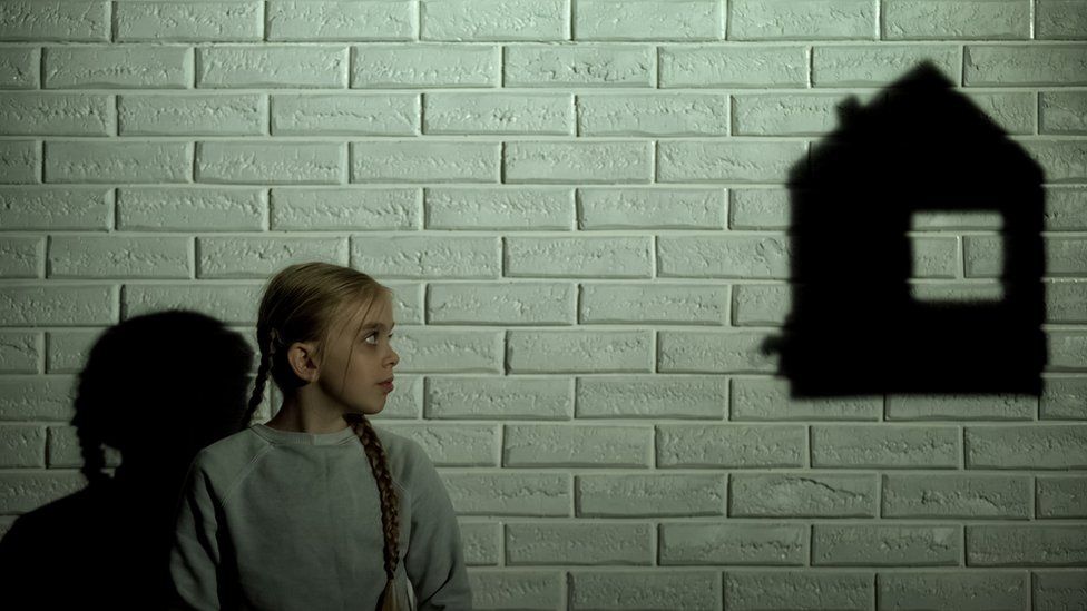 Young girl stood against white brick wall, looking towards a shadow representing a house