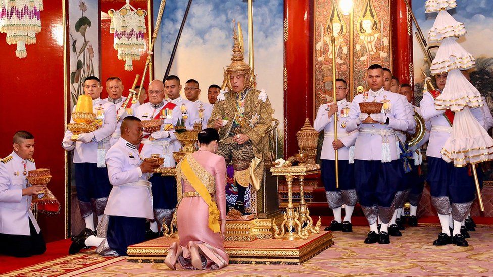 King Maha Vajiralongkorn wearing a gold crown while Queen Suthida kneels on the floor in front of him