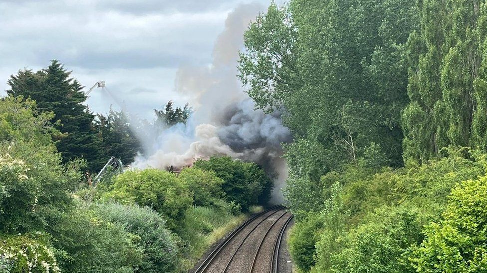 Trees and bushes either side of train tracks with thick black smoke billowing across
