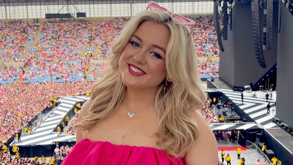 Lily Redman at a Harry Styles gig. Lily is a blonde woman in her 20s with wavy hair down past her shoulders. Lily is smiling at the camera and wears pink heart-shaped sunglasses on her head , bright pink lipstick and pink eye shadow embellished with small silver gems around her eyes. Behind her is the stage with a black and white striped floor and crowds of fans starting to gather in their seats