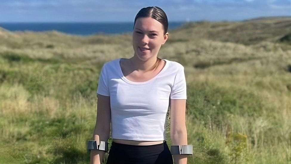 Jade Brooks standing on clifftop grassland, wearing a white top and a black trousers, in crutches 