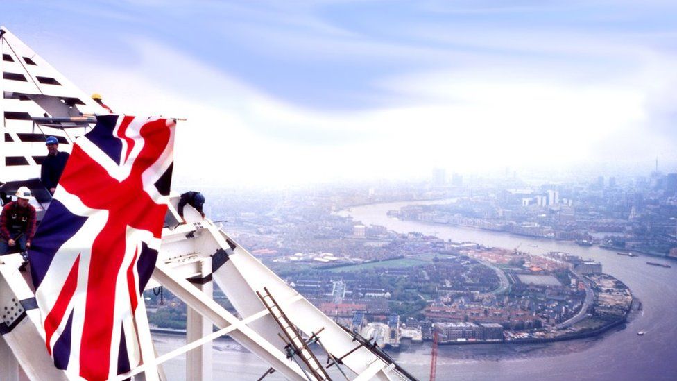 A photograph showing steel workers attaching the pyramid top to the skyscraper, with the River Thames and city behind.