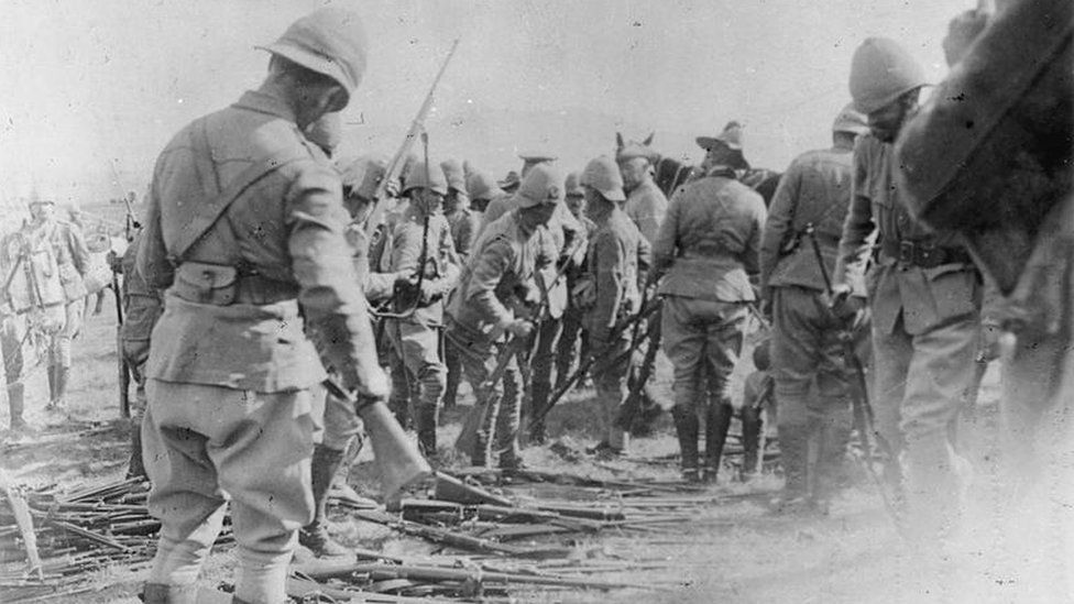 A faded black and white photograph showing a group of soldiers standing around a cache of rifles lying on the floor