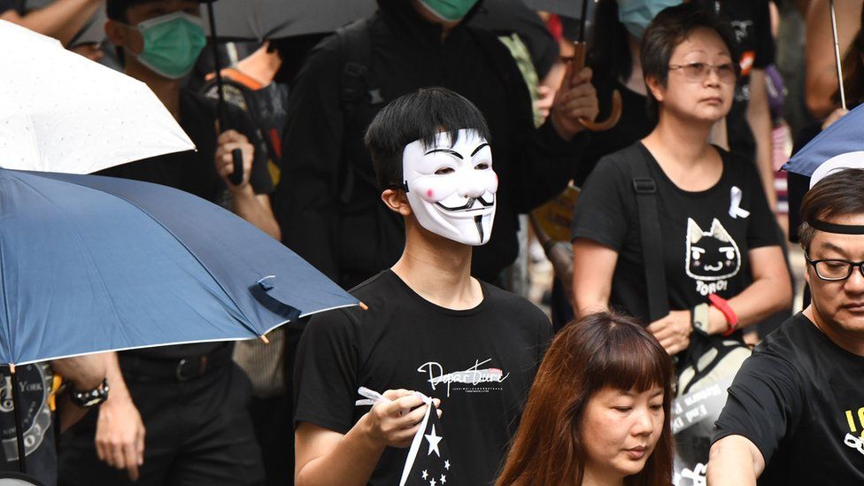 A protester (C), seen wearing a Guy Fawkes mask and holding a representation of the Chinese national flag coloured black,