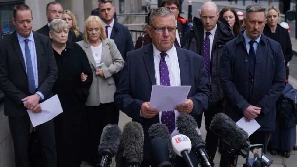 Gary Furlong - a white man wearing a dark suit and purple tie, with grey hair and glasses - speaking to the media after the inquest's conclusion. The victims' families and friends stand behind him, outside the Old Bailey in London