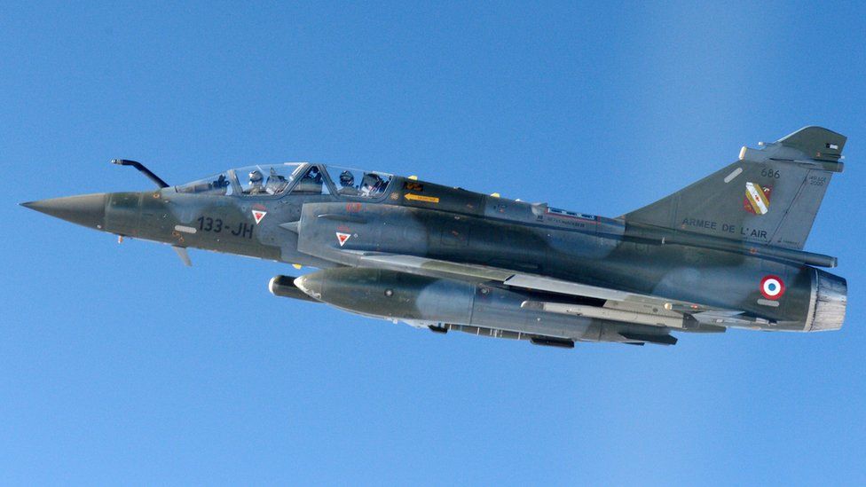 A Mirage 2000-D aircraft with a dark grey camouflage paint job is seen flying in a close-up shot against a bright blue sky