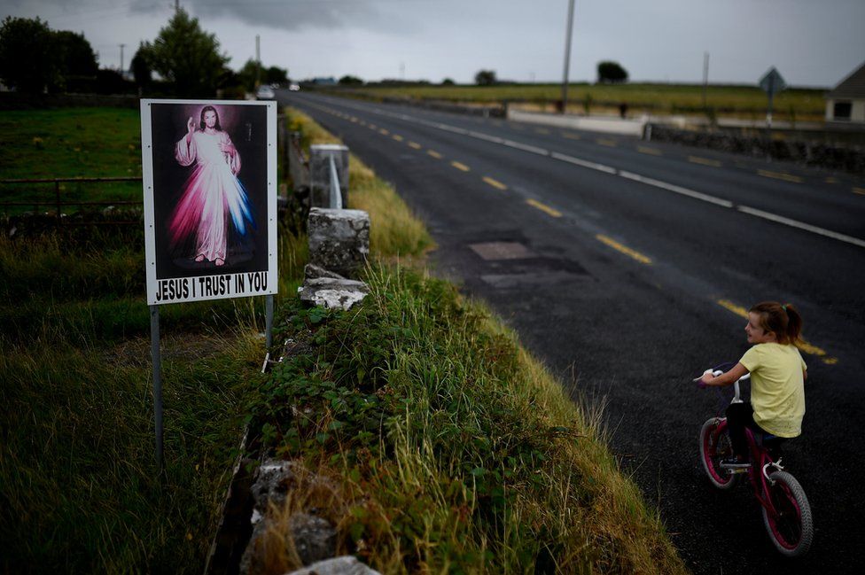 A child cycles past a roadside sign reading "Jesus I trust in you" near Tuam