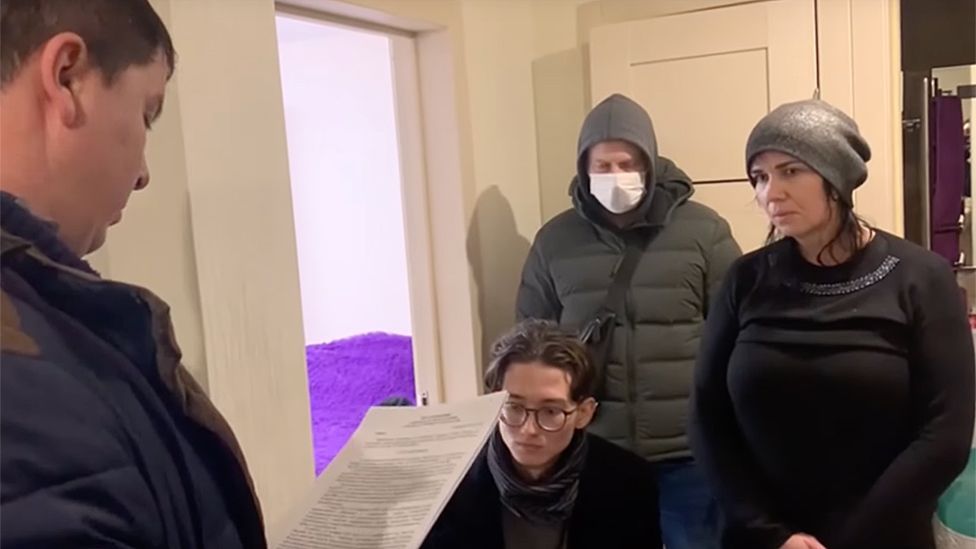 Kevin sitting down with his mother standing beside him, while one FSB officer looks at a piece of paper and another officer stands behind Kevin wearing a medical mask and hooded jacket