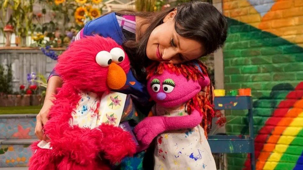 An actress in Sesame Street hugs the muppets Elmo and Lily