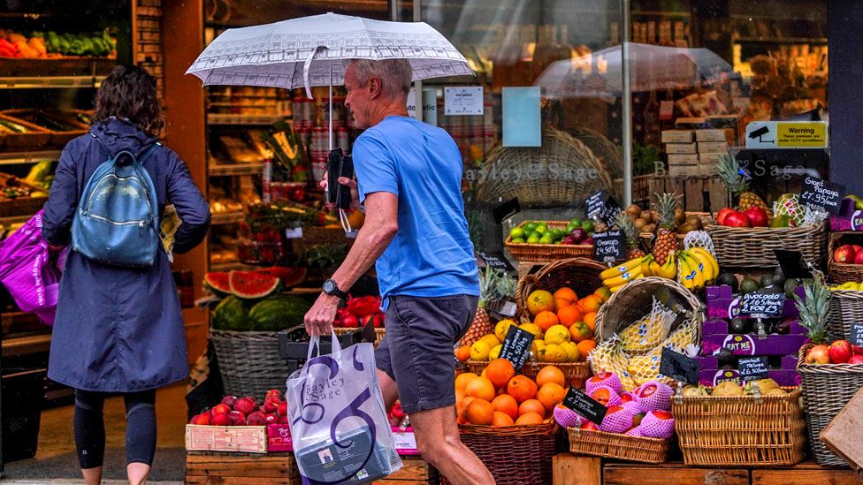 A shopper shelters under an umbrella in Wimbledon high street, south west London, during rain showers on 22 July 2023