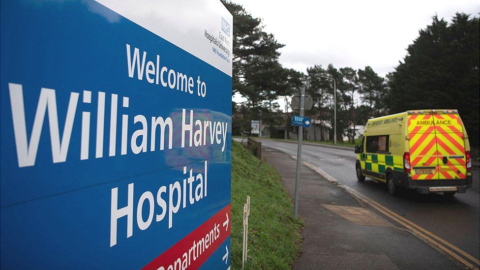 Signpost: "Welcome to William Harvey Hospital"; an ambulance drives by in the background