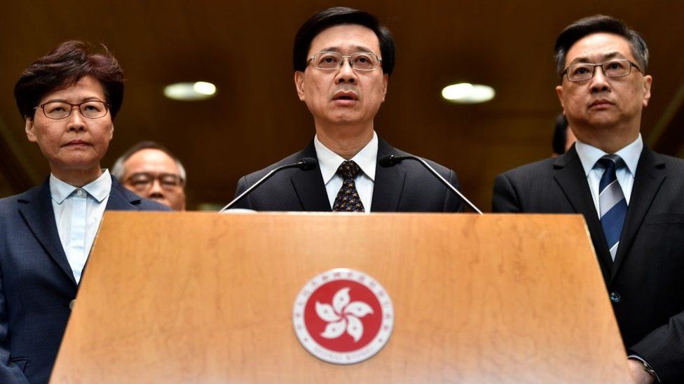 Глава исполнительной власти Гонконга Кэрри Лам (слева) и начальник полиции Стивен Ло (справа) наблюдают за тем, как глава службы безопасности Гонконга Джон Ли (в центре) обращается к СМИ на пресс-конференции в Гонконге 22 июля 2019 года.