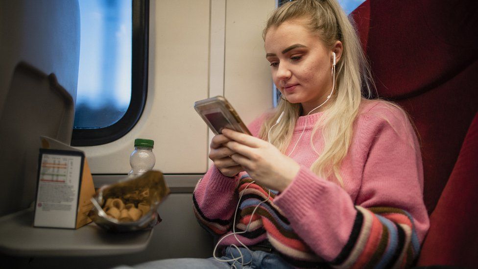 A young woman sat on a train with headphones on, looking at her phone