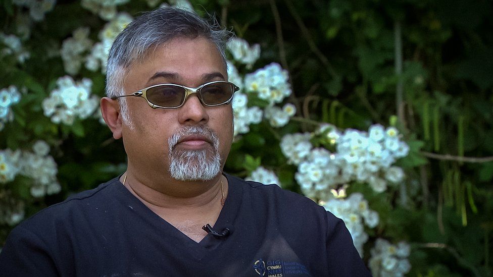 Mohan De Silva in a medical top and sunglasses sitting in a garden in front of white flowers